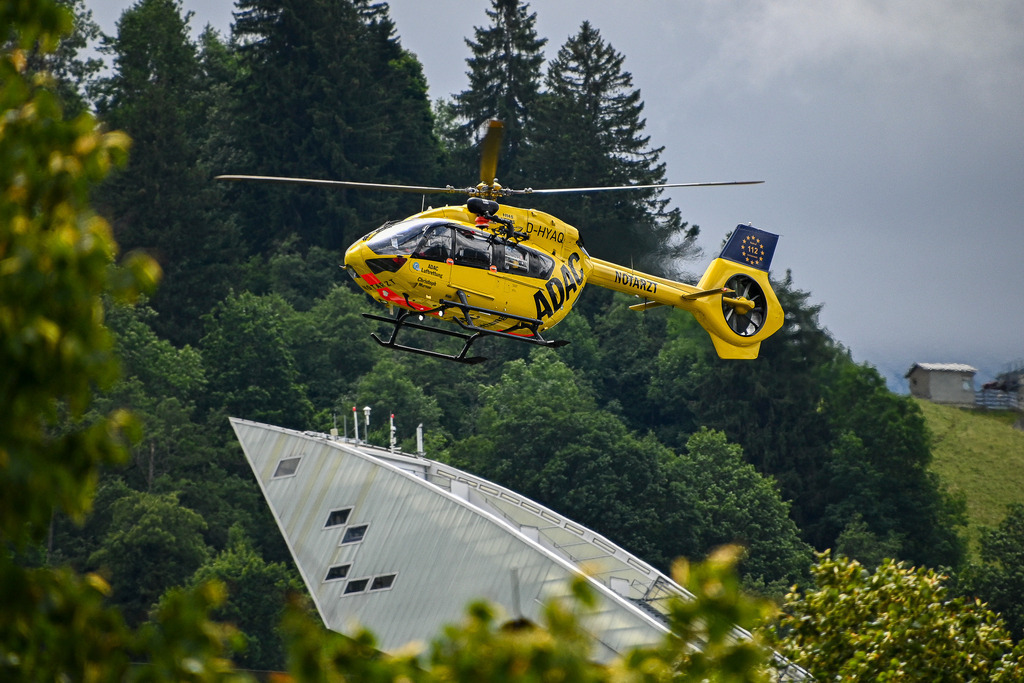 Retter aus der Luft: Hoffnung über der Olympia-Skischanze | Ein leuchtend gelber ADAC-Rettungshubschrauber schwebt in dynamischer Schräglage unmittelbar über der markanten, futuristischen Spitze der Olympiaschanze in Garmisch-Partenkirchen. Im Hintergrund erhebt sich die dicht bewaldete, alpine Bergkulisse. Das Zusammenspiel aus ikonischer Sportarchitektur und der Präzision des Helikopters im Flug vermittelt ein tiefes Gefühl von Einsatzbereitschaft und Sicherheit. Ein kraftvolles Bild, das die Gratwanderung zwischen Nervenkitzel im Leistungssport und der beruhigenden Gewissheit schneller medizinischer Hilfe im alpinen Raum symbolisiert. - Realisiert mit Pictrs.com