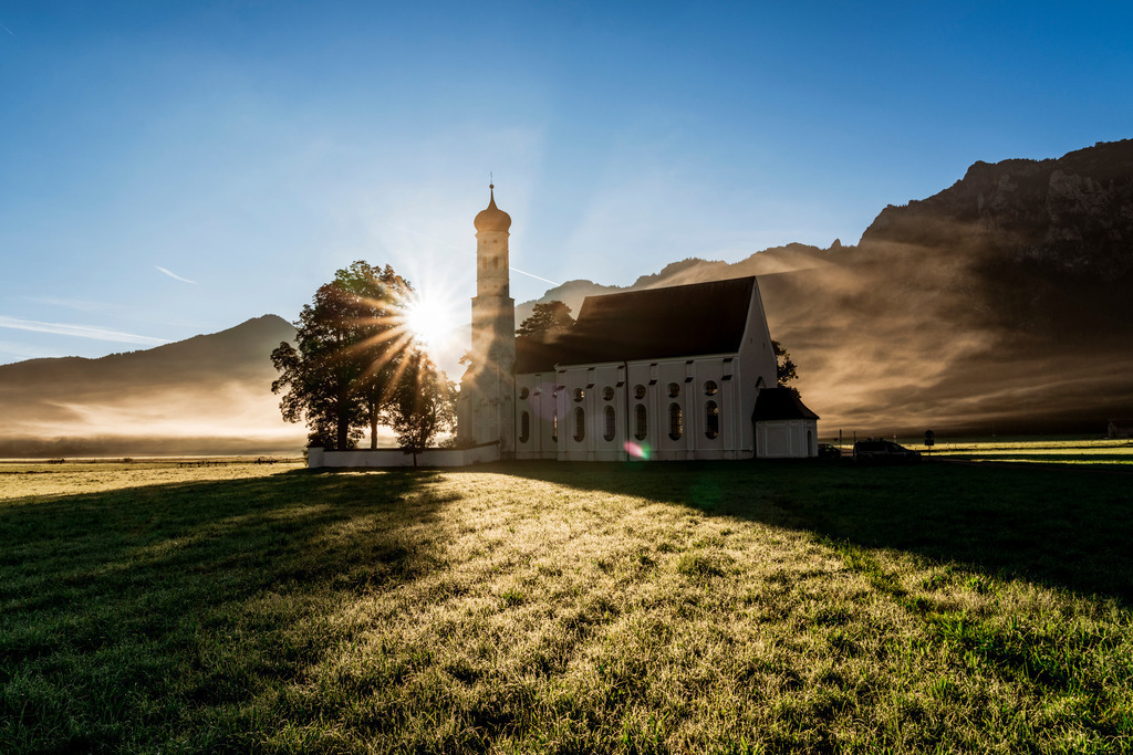 Allgäu Wandbild -  St. Coloman im Sonnenaufgang | Dieses Bild markiert in meinen Augen den Beginn des Herbsts in unserem schönen Fleckchen Allgäu. Der erste Bodennebel wird von der Sonne, welche wieder hinter den Bergen aufgeht, in ein wunderbar warmes Licht getaucht.