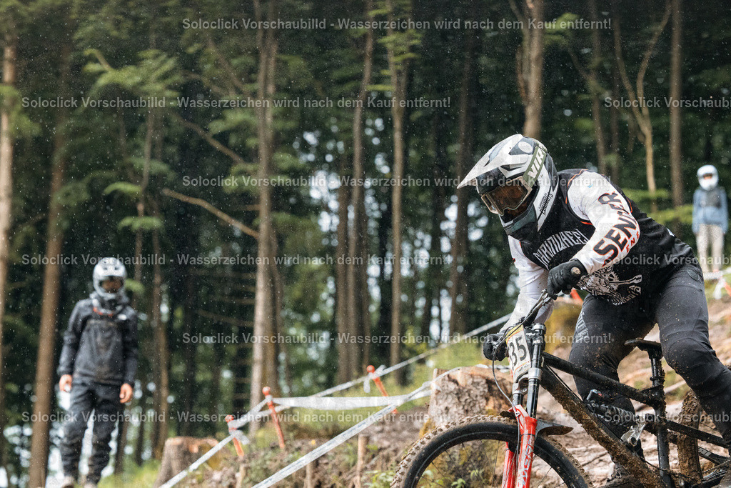 Mountainbike-Abenteuer im Regen | Ein Mountainbiker rast einen steilen, schlammigen Pfad hinunter, umgeben von dichtem Grün und Bäumen. Der Fahrer trägt einen schwarzen Helm und einen schwarzen Suit mit dem Schriftzug 'SENRA'. Er fährt ein Mountainbike mit roten Akzenten. Im Hintergrund sind weitere Personen in Schutzkleidung zu sehen, die den Fahrer beobachten. Es regnet leicht, was die Strecke rutschig macht. - Realisiert mit Pictrs.com