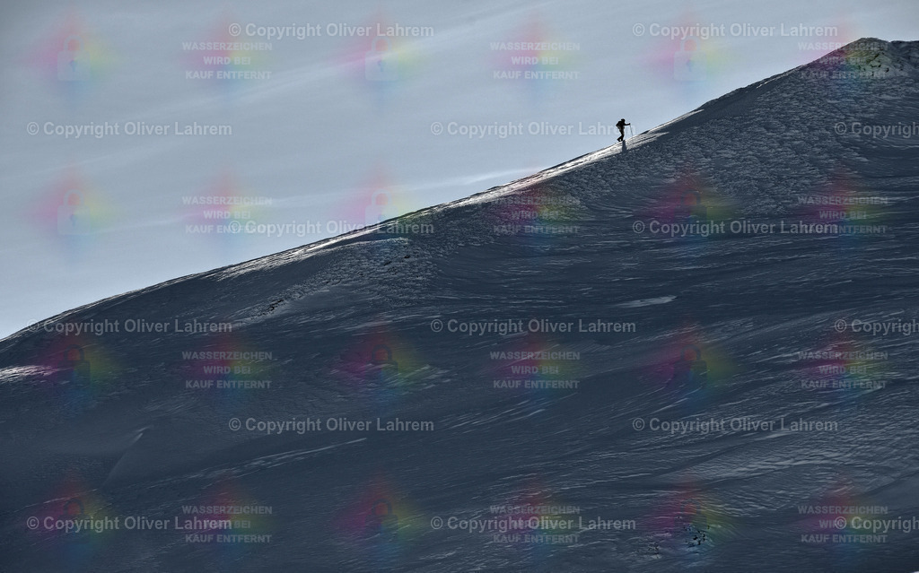 Kurz vor dem Gipfel | Ein Skitourengeher bei Anstieg an einem Berg bei blauem Himmel und der Wind wirbelt den leichten Pulverschnee auf.