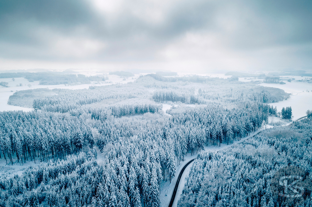 Winterlandschaft Oberschwaben – Verschneiter Wald und Straße aus der Luft | Diese Drohnenaufnahme zeigt eine weitläufige Winterlandschaft in Oberschwaben. Eine geschwungene Straße führt durch dichten, schneebedeckten Nadelwald und öffnet sich zu verschneiten Feldern. Die Luftperspektive offenbart die sanften Hügel und die klare Struktur der Landschaft unter bewölktem Winterhimmel. - Realisiert mit Pictrs.com