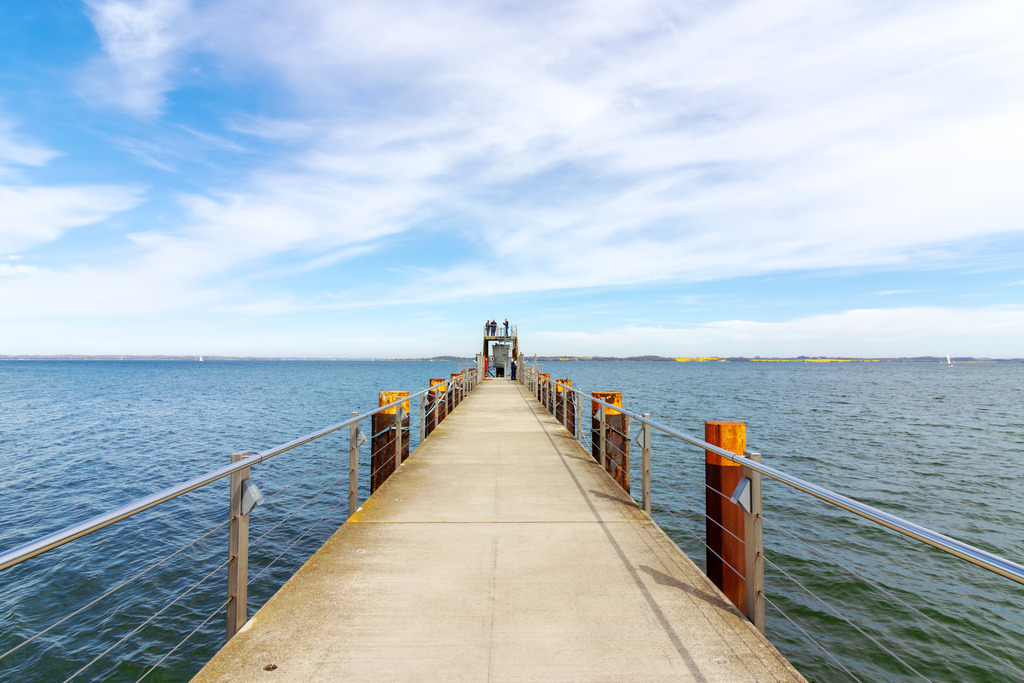 Wandbild: Küstenbrücke – Der Weg zur Förde-Weite | Genießen Sie den freien Blick über die Flensburger Förde bis hin zum dänischen Ufer. Dieses maritime Wandbild besticht durch seine beeindruckende Tiefenwirkung und die klare, nordische Lichtstimmung. Die Seebrücke in Langballigau führt den Blick unter einem dramatisch-schönen Wolkenhimmel direkt auf das blau schimmernde Wasser. Ein Motiv voller Freiheit und sommerlicher Frische, das Ihren Wohnraum optisch öffnet und die Sehnsucht nach dem Meer jeden Tag aufs Neue stillt. - Realisiert mit Pictrs.com