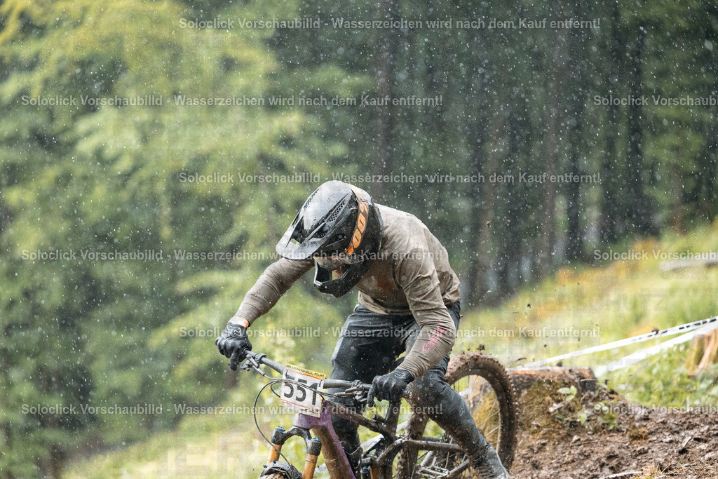Mountainbiker im Regen | Ein Mountainbiker in voller Ausrüstung rast durch einen verschneiten Wald. Er trägt einen schwarzen Helm mit einer gelben Visier und eine schwarze Maske. Sein Trikot ist schlammbedeckt. Er fährt ein violett-blaues Mountainbike mit schwarzen Reifen. Der Hintergrund besteht aus dichtem Grün und braunen Bäumen. Es regnet. - Realisiert mit Pictrs.com
