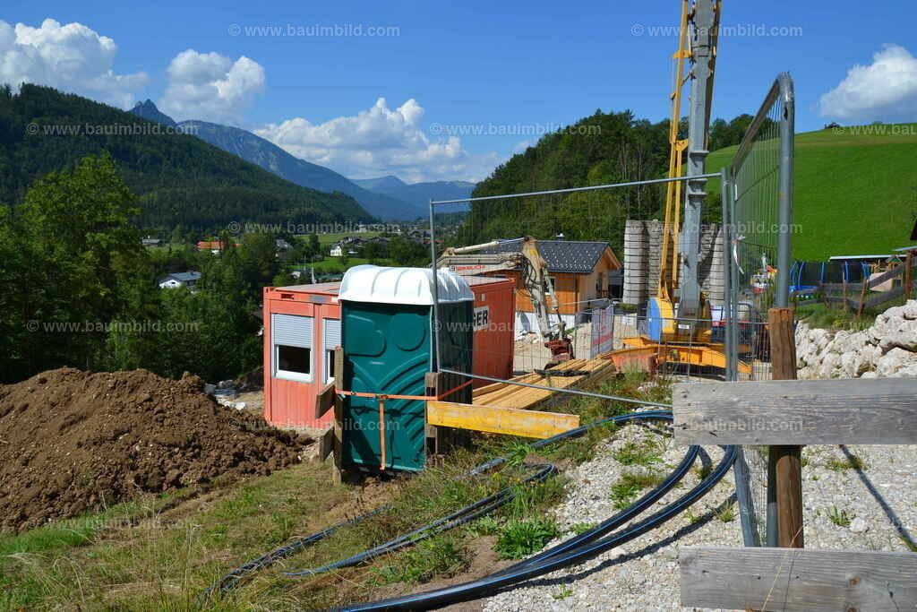 Baustelleneinrichtung | Roter Baucontainer, grünes Bau-WC, Baukran, Bagger, Absperrgitter und Holzzaun zur Absicherung der Baustelle, Berglandschaft, blauer Himmel mit Wolken
