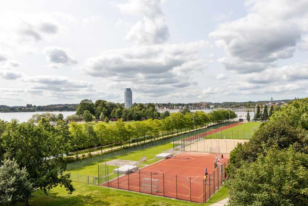Wandbild: Ausblick auf die Königswiesen in Schleswig | Dieses Wandbild im Querformat zeigt den Ausblick auf die Königswiesen vom Aussichtsturm in Schleswig. In der Ferne kann man den Wikingturm sehen. In der Nähe befinden sich Sportanlagen. - Realisiert mit Pictrs.com