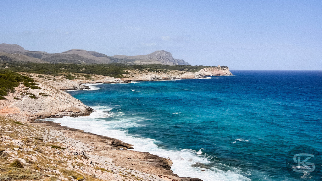 Spektakuläre Steilküste und Panoramablick bei Cala Mesquida, Mallorca | Panorama-Aufnahme einer felsigen Steilküste auf Mallorca bei Cala Mesquida mit schäumender Brandung am Ufer, tiefblauem Meer und einer weitläufigen Hügellandschaft unter klarem Himmel. - Realisiert mit Pictrs.com