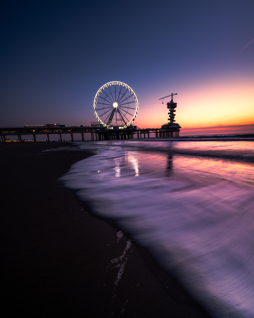Riesenrad von Scheveningen zur blauen Stunde | Das beleuchtete Riesenrad auf dem Pier von Scheveningen spiegelt sich während der blauen Stunde im nassen Sand des Strandes. - Realisiert mit Pictrs.com