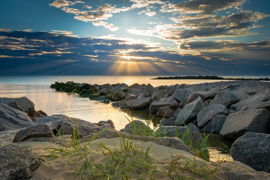 Steinbuhnen am Schönberger Strand | Das Bild zeigt Steinbuhnen am Schönberger Strand. Der Himmel ist leicht bewölkt und einzelne Sonnenstrahlen durchbrechen die Wolkendecke. Diese tauchen die Szenerie in ein warmes, goldenes Licht.
Die Steinbuhne wirkt wie eine natürliche Barriere gegen das aufgewühlte Meer, während das Rauschen der Wellen die Luft erfüllt und eine entspannende Atmosphäre schafft. Die sanfte Brise des Meeres bewegt das Gras und lässt die Schönheit der Umgebung noch intensiver erleben.
Im Hintergrund ist der Horizont zu sehen, wo sich das Meer und der Himmel miteinander verbinden und eine unendliche Weite erschaffen. Eine friedliche Stimmung liegt in der Luft und lässt den Betrachter den Moment genießen und in die Schönheit der Natur eintauchen.