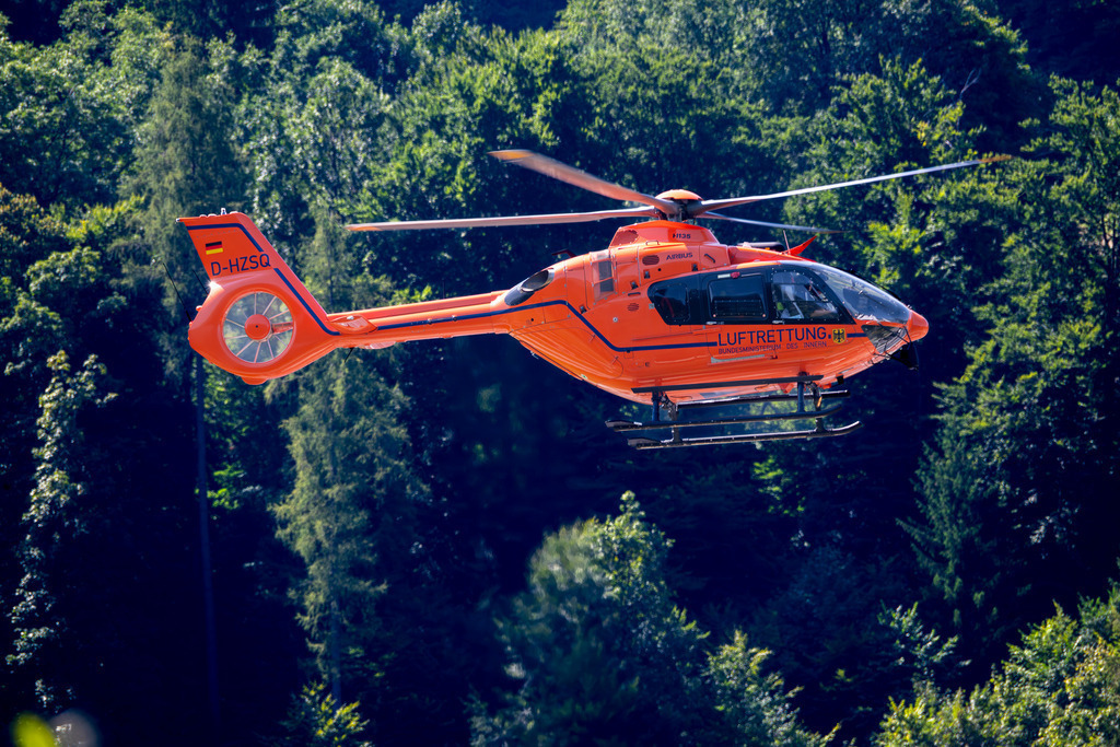 Rettungshubschrauber | Ein leuchtend oranger Rettungshubschrauber der Zivilschutz-Luftrettung (D-HZSQ) schwebt majestätisch über einem dichten, dunkelgrünen Nadelwald. Die Sonne bringt die Farbe der Maschine zum Leuchten, während die sanften Hügel im Hintergrund in ein sommerliches Licht getaucht sind. - Realisiert mit Pictrs.com