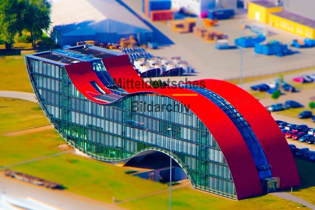 Luftbild Magdeburg Enercon-Welle Miniatur IMG_2209 | Das Luftbild zeigt das markante Bürogebäude des Windenergieanlagenherstellers Enercon in Magdeburg aus einer Drohnenperspektive. Die geschwungene Gebäudeform erinnert an eine Welle und hebt sich mit ihrer modernen Architektur deutlich von den umliegenden Bauwerken ab. Aus der Vogelperspektive wird die außergewöhnliche Linienführung besonders eindrucksvoll sichtbar und unterstreicht den innovativen Charakter des Unternehmens.Das Verwaltungsgebäude wurde als moderner Bürokomplex für den Enercon-Standort Magdeburg errichtet. Die geschwungene Fassade greift die Dynamik der Windenergie auf und spiegelt die technische Ausrichtung des Unternehmens wider. Am Standort Magdeburg werden unter anderem Komponenten für Windenergieanlagen entwickelt und gefertigt. Enercon zählt mit mehr als 30.000 errichteten Windenergieanlagen weltweit zu den führenden Herstellern in diesem Bereich und setzt seit Jahrzehnten auf getriebelose Antriebstechnik als besonderes Konstruktionsmerkmal.Die Drohnenaufnahme verdeutlicht die harmonische Verbindung von moderner Architektur und Ingenieurwesen. Die außergewöhnliche Gebäudeform macht den Enercon-Standort zu einem markanten Beispiel zeitgenössischer Industriearchitektur und unterstreicht die Bedeutung Magdeburgs als Standort für innovative Energie- und Umwelttechnik. - Realisiert mit Pictrs.com