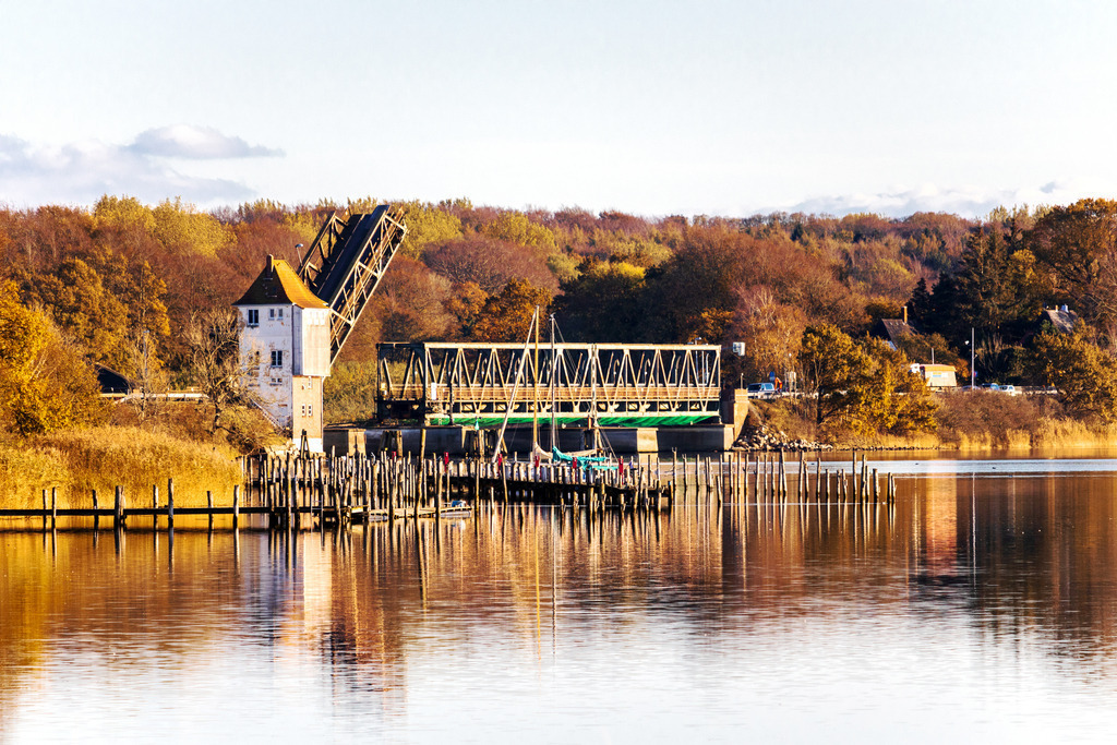 XXL Wandbild Historische Schleibrücke im goldenen Herbst – Leinwandbild | Technisches Denkmal und herbstliche Idylle im Querformat. Dieses XXL Wandbild zeigt die markante Lindaunisbrücke, die sich majestätisch über den Ostseefjord Schlei spannt. Das warme Licht des goldenen Oktobers lässt die umliegenden Wälder in kräftigen Braun- und Goldtönen leuchten, während sich die filigrane Stahlkonstruktion und der historische Brückenturm im spiegelglatten Wasser reflektieren. Holen Sie sich dieses Stück Heimatgeschichte nach Hause, um in Wohnzimmern oder Arbeitsbereichen einen Blickfang mit regionalem Charakter zu schaffen. Auch für Büros oder öffentliche Gebäude in der Region Angeln und Schwansen ist dieses Motiv eine hervorragende Wahl, um die Verbundenheit zur Schlei-Region auszudrücken. Die Kombination aus technischer Präzision und der sanften, herbstlichen Naturkulisse verleiht jedem Raum eine ruhige und zugleich interessante Ausstrahlung. Der Druck erfolgt auf hochwertiges, 260 g/m² schweres Leinwandgewebe. Die sorgfältige Aufspannung in Handarbeit auf einen 2 cm starken Holzkeilrahmen aus nachhaltiger Forstwirtschaft unterstützt eine dauerhafte Formstabilität und garantiert eine hochwertige Präsentation dieses zeitlosen Motivs. - Realisiert mit Pictrs.com