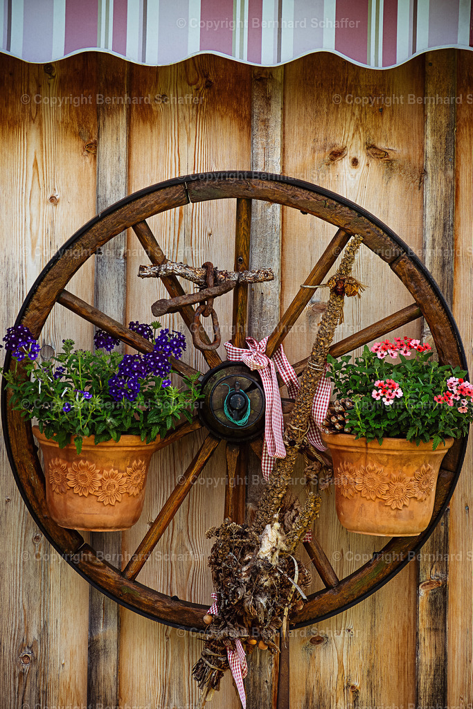 Wagenrad an der Wand | An einer Holzwand hängt ein mit Blumen geschmücktes Wagenrad mit Speichen. - Realisiert mit Pictrs.com
