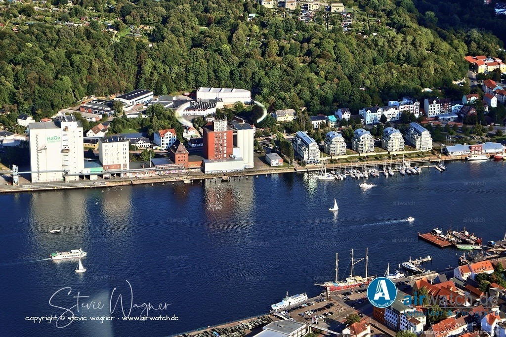 Luftbild Flensburg - Der Ballastkai-Speicher in Flensburg-Fruerlund | Der Ballastkai-Speicher in Flensburg-Fruerlund liegt unmittelbar an der Wasserkante des Flensburger Hafens und gehört zu den Kulturdenkmalen der Stadt. Der älteste Teil des Silokomplexes, ein dreißig Meter hoher, fünfgeschossiger Getreidespeicher, wurde 1936 nach Plänen des Berliner Oberingenieurs Max Schulz errichtet und im Heimatschutzstil gestaltet, wobei der Eisenbetonbau eine rote Klinkerverblendung erhielt. Auf der Nordseite entstand zeitgleich ein dreigeschossiger Anbau, der heute noch erhalten ist.