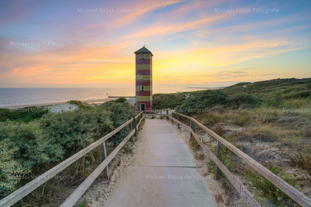 Leuchtturm in den Dünen bei Dishoek in Zeeland | Ein Weg führt durch die Dünen von Dishoek direkt auf den markanten Leuchtturm zu. Der untere Leuchtturm steht eindrucksvoll im Mittelpunkt des Bildes und hebt sich klar von der weiten Dünenlandschaft ab.Die Sonne ist bereits untergegangen, doch der Himmel leuchtet noch in intensiven Gelb-, Orange- und Rottönen. Dieses warme Nachglühen bildet einen eindrucksvollen Kontrast zum dunkler werdenden Leuchtturm und den Silhouetten der Dünen.Ein stimmungsvolles Landschaftsfoto aus Dishoek in Zeeland, Niederlande, das die besondere Atmosphäre der blauen Stunde kurz nach Sonnenuntergang einfängt. - Realisiert mit Pictrs.com