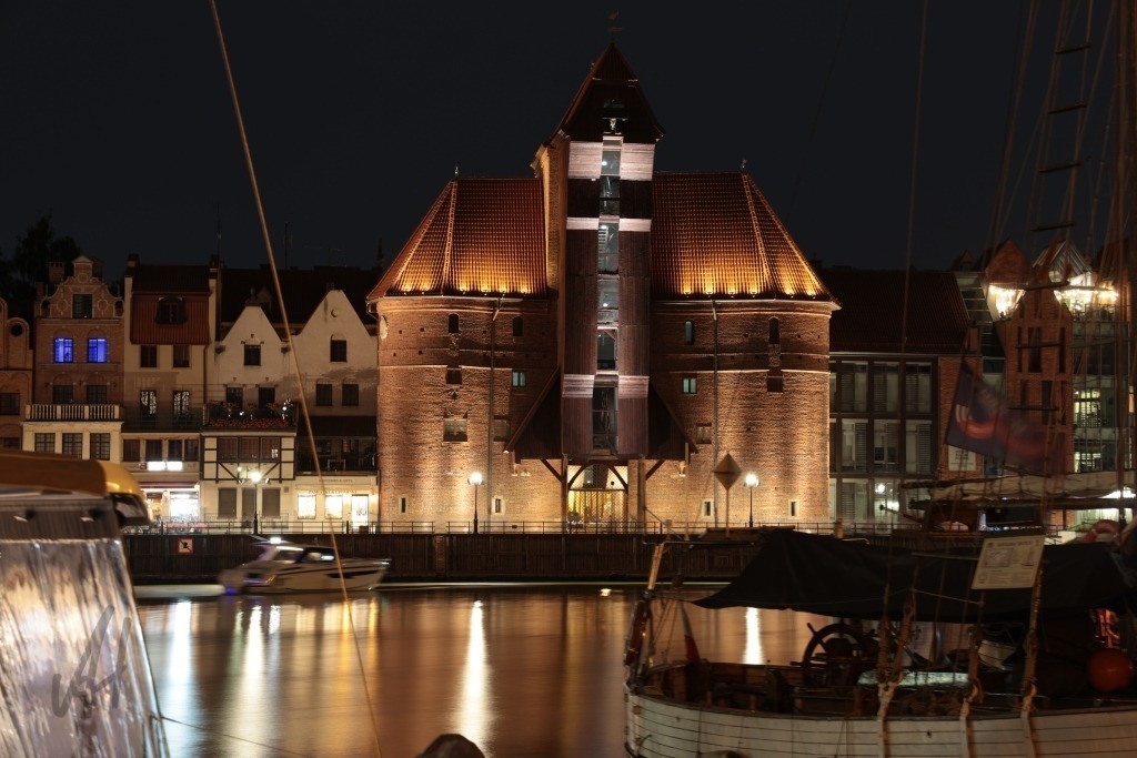 Krantor Danzig bei Nacht | Das Foto zeigt das Krantor von Danzig (Gdańsk) bei Nacht, eines der bekanntesten Wahrzeichen der historischen Hansestadt. Das mächtige mittelalterliche Hafentor erhebt sich eindrucksvoll über der Mottlau und wird von der nächtlichen Beleuchtung wirkungsvoll in Szene gesetzt. Die warmen Lichter spiegeln sich auf der dunklen Wasseroberfläche und erzeugen ein stimmungsvolles Farbenspiel. Rund um das Krantor sind die historischen Fassaden der Uferbebauung zu erkennen, deren beleuchtete Fenster und Konturen die maritime Atmosphäre verstärken. Die ruhige Mottlau bildet dabei einen natürlichen Vordergrund und verleiht dem Bild zusätzliche Tiefe. Besonders eindrucksvoll ist der Kontrast zwischen dem dunklen Nachthimmel, den warm beleuchteten historischen Gebäuden und den Lichtreflexen im Wasser. Das Foto vermittelt eine ruhige, romantische und zugleich geschichtsträchtige Atmosphäre und lässt die jahrhundertealte Bedeutung Danzigs als bedeutende Hafen- und Handelsstadt erahnen. - Realisiert mit Pictrs.com