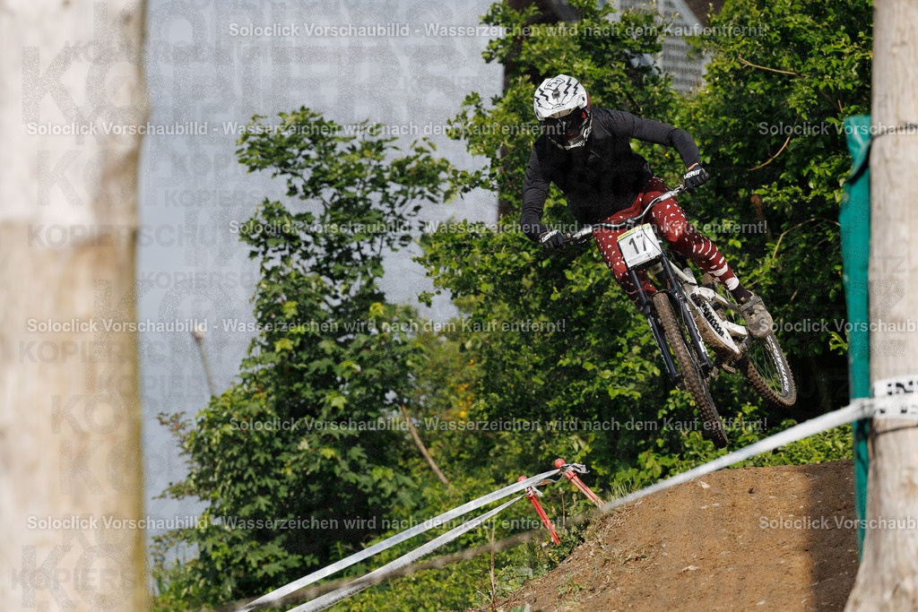 Mountainbiker im Flug | Ein Mountainbiker ist in voller Fahrt auf einem staubigen Trail zu sehen. Er macht einen Sprung und scheint die Kontrolle über sein Fahrrad zu haben. Der Hintergrund besteht aus dichtem Grün und einem Blick auf das Meer. - Realisiert mit Pictrs.com