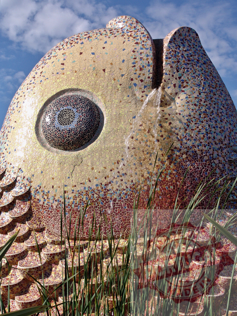 Colorful mosaic fish fountain spraying water | Mosaic fish sculpture with vibrant tiles forming scales, spraying water under a blue sky - Realized with Pictrs.com