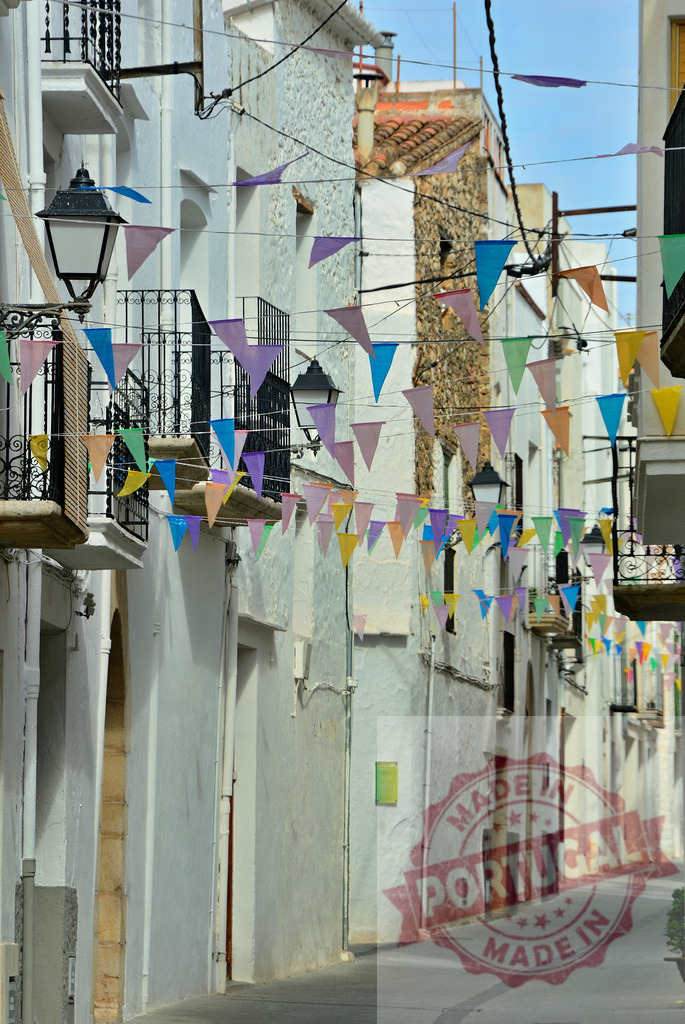 Spanish village street with colorful celebration bunting. Spain - Castellon | Narrow street of white buildings decorated with vibrant pennant flags, creating a festive scene - Realized with Pictrs.com