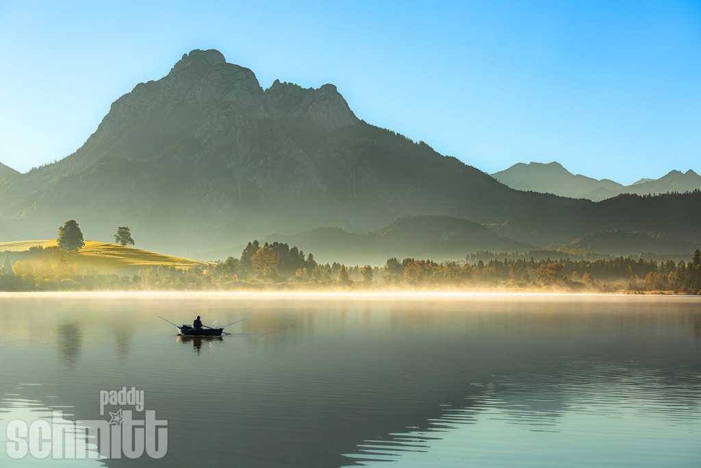 Angler auf dem Hopfensee vor dem Säuling im Morgennebel | Ein Angler auf dem ruhigen Hopfensee vor der markanten Silhouette des Säulings, umgeben von sanftem Morgennebel. Eine stimmungsvolle Aufnahme, die die besondere Ruhe und Atmosphäre des frühen Morgens einfängt. - Realisiert mit Pictrs.com