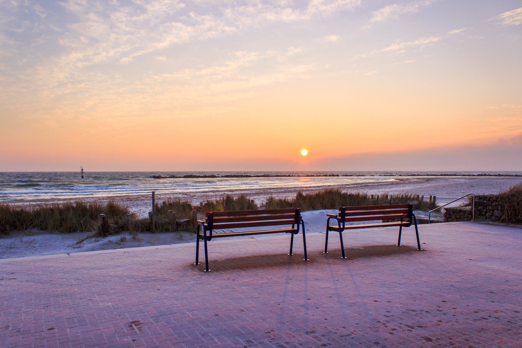 Wandbild: Parkbänke im Sonnenaufgang an der Ostsee | Dieses Wandbild im Querformat zeigt eine schöne maritime Morgenstimmung. Im Vordergrund befinden sich zwei Parkbänke im Licht der aufgehenden Sonne. Der Himmel leuchtet in einem schönen warmen Orangeton.  - Realisiert mit Pictrs.com