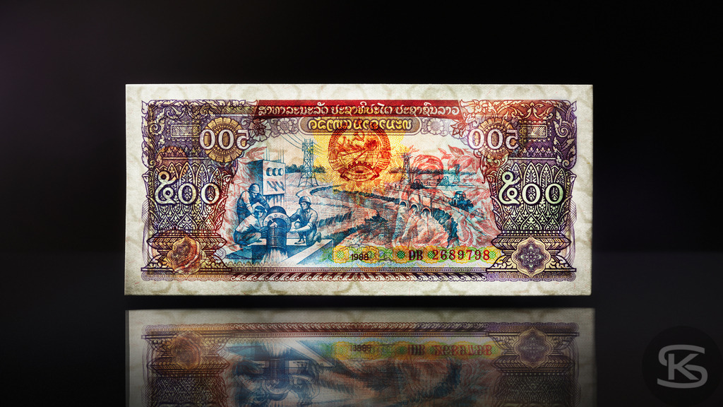 Laos 200 Kip Banknote 1988 – Laotische Währung & Geschichte | Detailaufnahme der Vorderseite einer laotischen 200-Kip-Banknote von 1988. Das Motiv zeigt Arbeiter bei der Landwirtschaft oder in einer Fabrik sowie Fahrzeuge. - Realisiert mit Pictrs.com