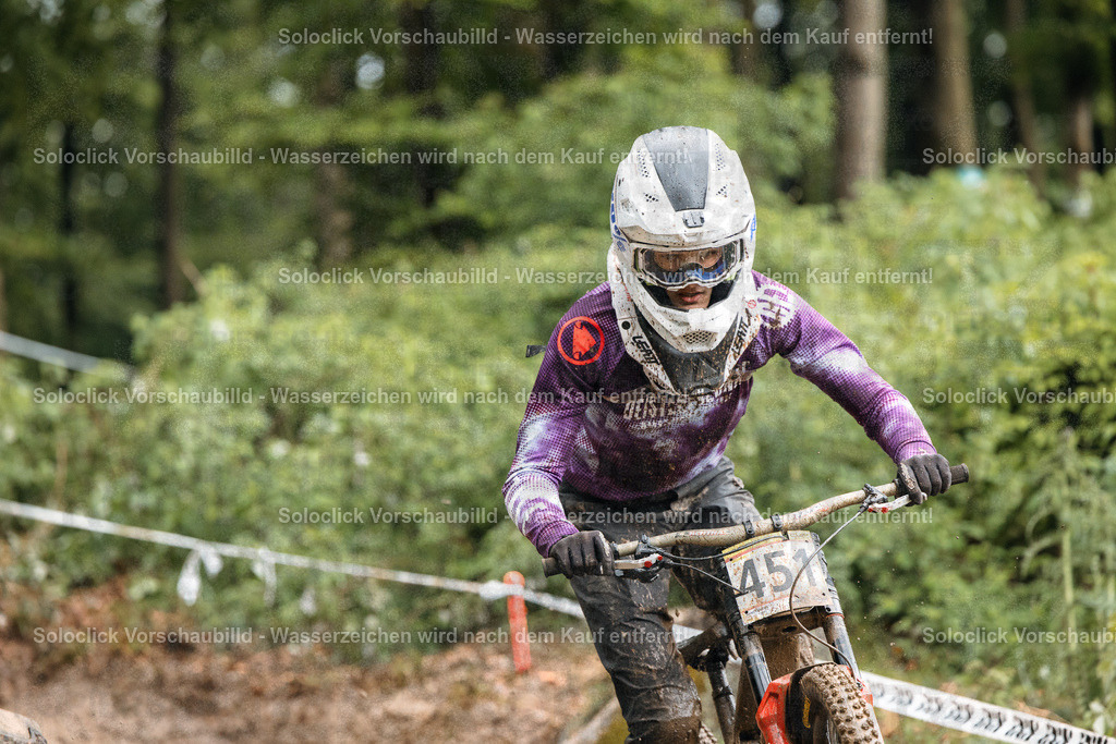 Mountainbiker im Sturmlauf | Ein Mountainbiker in voller Ausrüstung rast einen staubigen, kurvigen Trail hinunter. Er trägt einen weißen Helm mit dunklen Akzenten und eine violette Jacke mit dem Schriftzug 'REIGN'. Er fährt ein Mountainbike mit einem Nummernschild mit der Nummer 457. - Realisiert mit Pictrs.com