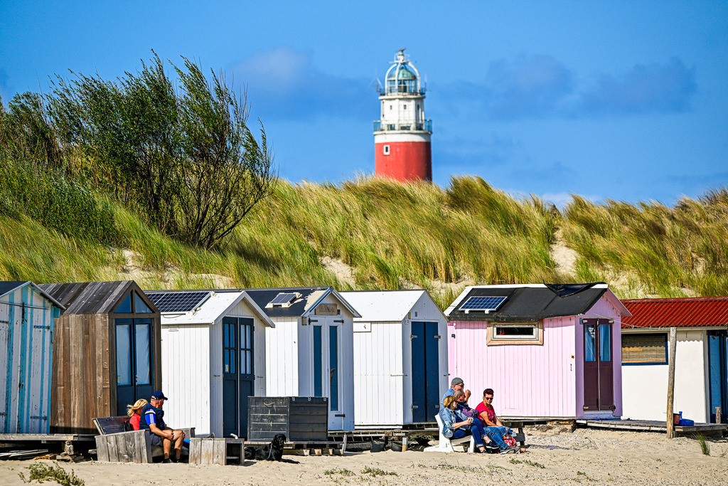Inselglück im Schatten des Leuchtturms | Vor der malerischen Dünenkulisse von Texel spiegeln bunt aufgereihte Strandhäuschen pure nordische Lebensfreude wider. Während Menschen und ihr Vierbeiner entspannt die warme Sonne genießen, thront der leuchtend rote Leuchtturm wahrzeichenhaft über dem Dünengras. Ein unbeschwerter Küstenmoment voll Harmonie, sanftem Meereszauber und zeitloser Urlaubsidylle an der Nordsee. - Realisiert mit Pictrs.com