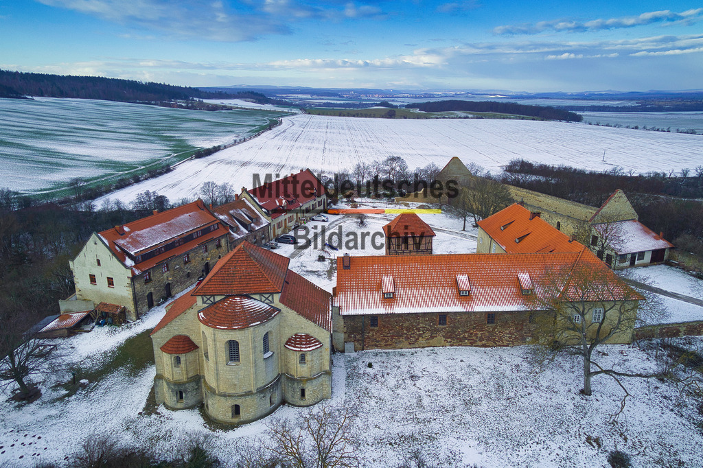 konradsburg_3_0030 | Die Aufnahme zeigt die Konradsburg bei Ermsleben in einer winterlichen Landschaft aus einer Drohnenperspektive. Eine leichte Schneedecke bedeckt die Dächer der historischen Gebäude und die umliegenden Felder und verleiht dem ehemaligen Burg- und Klosterkomplex eine ruhige, fast märchenhafte Atmosphäre. Im Vordergrund sind links die markante Bastei und rechts das ehemalige Wirtschaftsgebäude zu erkennen, während sich im Hintergrund die historischen Wohngebäude harmonisch in das Gesamtensemble einfügen.Die Konradsburg wurde im 11. Jahrhundert als hochmittelalterliche Burganlage der Grafen von Konradsburg errichtet. Bereits im Jahr 1120 wurde sie in ein Benediktinerkloster umgewandelt, dessen romanische Klosterkirche bis heute zu den bedeutendsten Sakralbauten der Region zählt. Nach der Auflösung des Klosters während der Reformation entwickelte sich die Anlage zu einem landwirtschaftlichen Gutshof. Die historischen Gebäude wurden im Laufe der Zeit erhalten und werden heute überwiegend als Wohngebäude genutzt.Die Drohnenaufnahme vermittelt einen eindrucksvollen Blick auf die außergewöhnliche Architektur und die geschlossene Anlage der Konradsburg. Die winterliche Landschaft unterstreicht den historischen Charakter dieses bedeutenden Kulturdenkmals und macht die wechselvolle Geschichte von Burg, Kloster und Gutshof aus einer einzigartigen Perspektive erlebbar. - Realisiert mit Pictrs.com