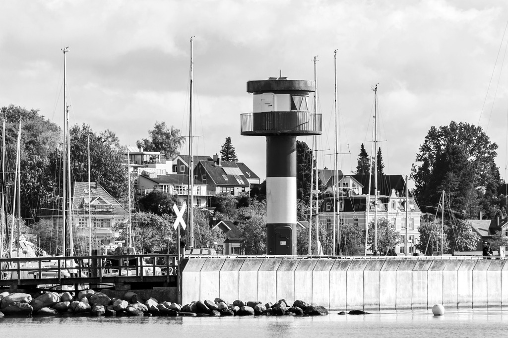 Wandbild: Leuchtturm in Eckernförde am Yachthafen in Schwarz-Weiß | Dieses Wandbild im Querformat zeigt den malerischen Leuchtturm am Yachthafen in Eckernförde in Schwarz-Weiß. Im Hintergrund sind die Masten der Segelboote sowie die einige Häuser zu sehen. - Realisiert mit Pictrs.com
