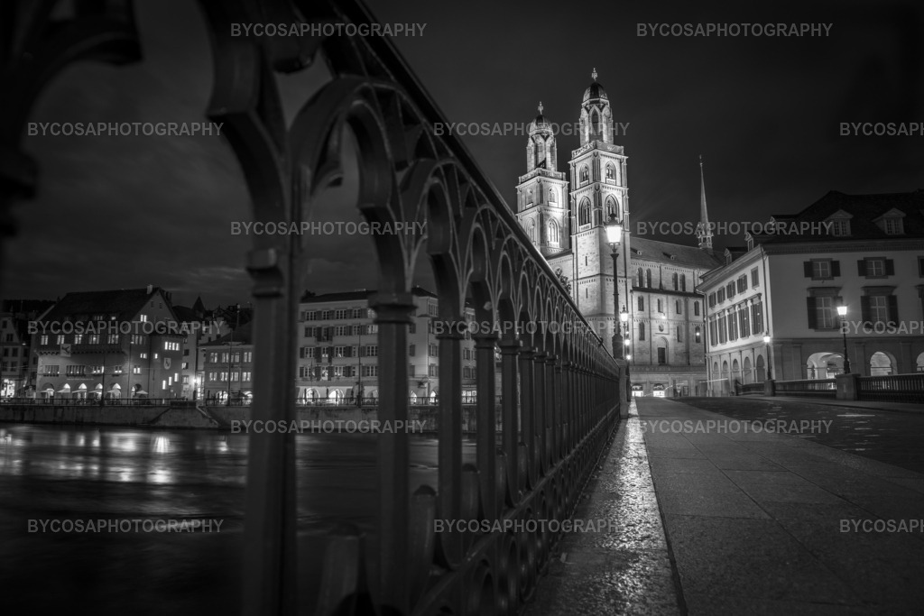 Night Path to Grossmünster _ Zürich | Eine eindrucksvolle Schwarz-Weiß-Fotografie in FineArt-Qualität, die das ikonische Grossmünster aus einer kraftvollen Froschperspektive bei Nacht einfängt. Die Linien des Metallgeländers lenken den Blick direkt auf die erleuchtete Kathedrale, während Spiegelungen auf dem Wasser und dem nassen Stein die Szene in eine elegante, friedvolle und perfekt ausbalancierte Nachtkomposition verwandeln. Ideal für moderne Wohnungen, luxuriöse Büros und Liebhaber von Minimalismus und Architektur. - Realisiert mit Pictrs.com