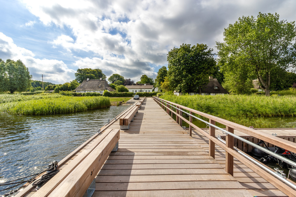 Wandbild: Steg in Sieseby an der Schlei | Dieses Wandbild im Querformat zeigt den neuen Steg in Sieseby an der Schlei im Sommer 2024. In der Ferne kann man die schönen Reetdachhäuser direkt am Schleiufer sehen. Im Uferbereich ist hellgrünes Schilf sowie schöne Bäume zu sehen. Am blauen Himmel befinden sich einige sommerliche Wolken. - Realisiert mit Pictrs.com