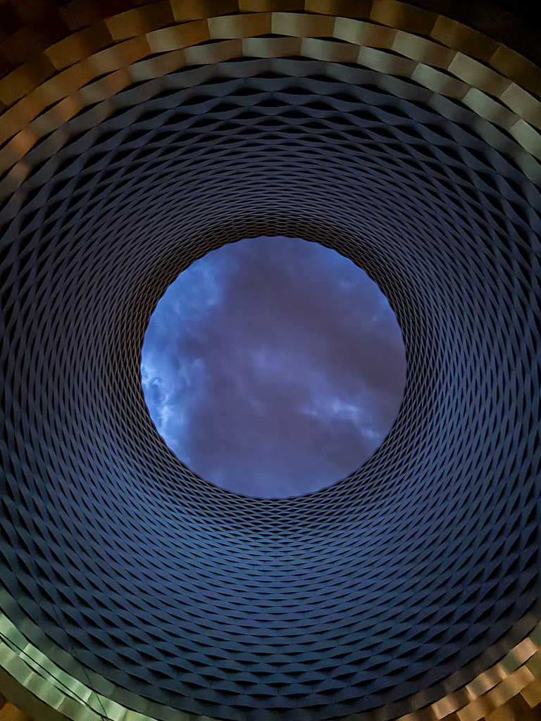 Wandbild Fenster zum Abendhimmel - Architektonischer Vortex | Dieses außergewöhnliche Fine-Art-Wandbild fasziniert durch eine spektakuläre optische Täuschung aus dem echten Leben. Der meisterhafte Blick durch eine spiralförmig geschwungene Dachfenster-Konstruktion führt das Auge direkt in den klaren Abendhimmel während der blauen Stunde. Was auf den ersten Blick wie ein Science-Fiction-Vortex oder ein ferner Planet im Weltall wirkt, ist das harmonische Zusammenspiel aus moderner Architektur-Geometrie und der Natur bei Dämmerung. Fotograf Robert Steinke fängt diese haptisch gemusterte Tunnel-Struktur und die beruhigende, kosmische Tiefe in gestochen scharfer Premium-Qualität ein. Ein absolut progressiver Blickfang für moderne Wohnzimmer, urbane Lofts oder repräsentative Büroräume. Perfekt als Kunstdruck auf Alu-Dibond oder unter brillantem Acrylglas für maximale Raumtiefe. - Realisiert mit Pictrs.com