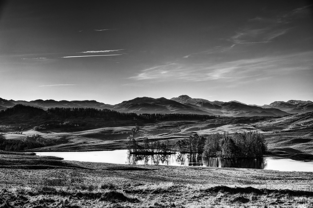 Loch Moraig | Eine weite, hügelige Landschaft in Schwarz-Weiss erstreckt sich unter einem klaren Himmel. Im Vordergrund liegt ein ruhiger See mit bewaldeten Inseln, deren Spiegelungen sich im Wasser abzeichnen. Die tief stehende Sonne erzeugt starke Kontraste und modelliert die sanften Formen der schottischen Highlands. - Realisiert mit Pictrs.com
