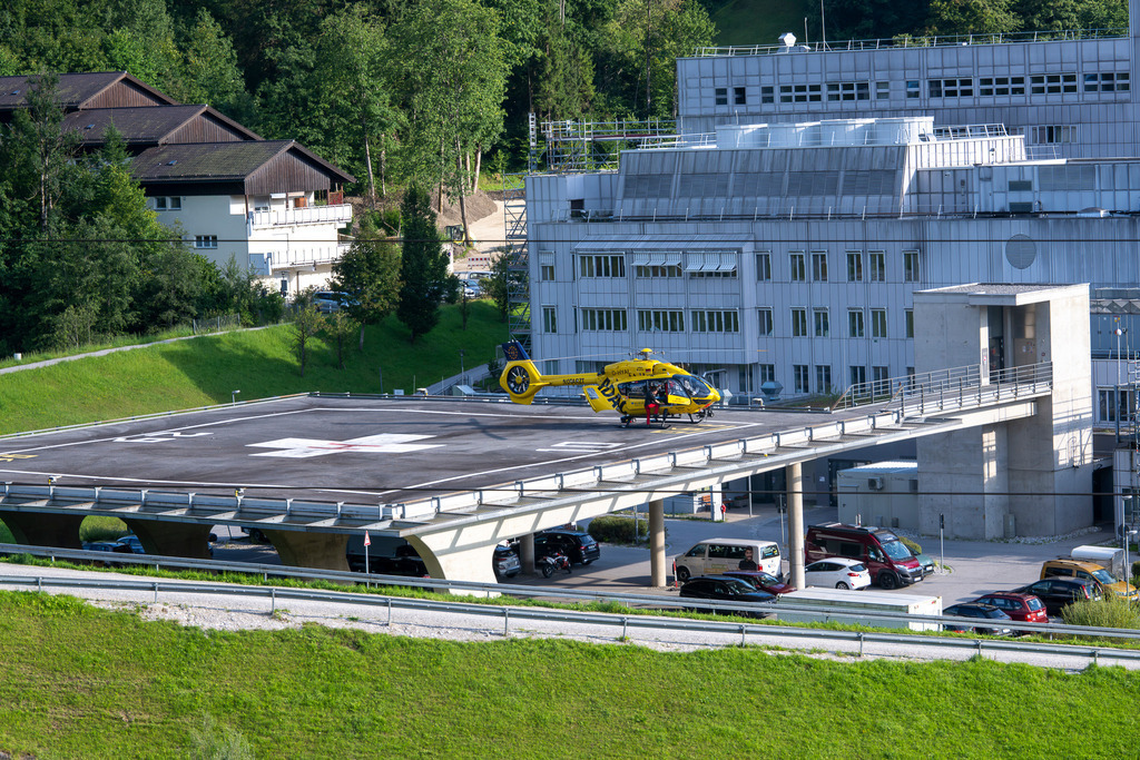 ADAC Rettungshubschrauber am Klinikum Garmisch-Partenkirchen | Ein markanter gelber ADAC-Rettungshubschrauber (Kennung D-HYAL) steht mit geöffneter Kabinentür auf dem modernen, erhöhten Landedeck des Klinikums Garmisch-Partenkirchen. Die Szene zeigt den Kontrast zwischen der funktionalen Krankenhausarchitektur, dem saftigen Grün der umliegenden Hänge und der technischen Präzision der Luftrettung in Warteposition. - Realisiert mit Pictrs.com