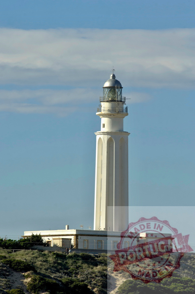 Faro de Trafalgar on coast guiding navigation. Espana - Cadiz | White lighthouse standing tall on a grassy dune, providing a beacon for ships - Realized with Pictrs.com