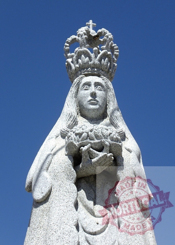 Virgin Mary stone statue with crown symbolizing faith. Spain - Extremadura | Virgin Mary statue carved from granite featuring a crown and holding flowers, against a blue sky - Realisiert mit Pictrs.com