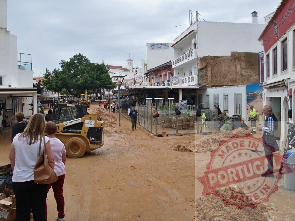 City street cleaning mud after flood disaster | Albufeira Flood - discruction by weather problems A bobcat and workers clearing mud from a city street after a flood - Realized with Pictrs.com