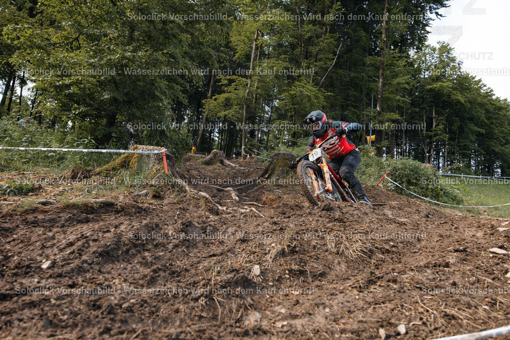 Mountainbike-Action im Gelände | Ein Mountainbiker in voller Ausrüstung, einschließlich Helm und Schutzbrille, navigiert mit hoher Geschwindigkeit durch eine anspruchsvolle Strecke. Der Boden ist staubig und wellig, was die Herausforderung des Geländes unterstreicht. Die Umgebung besteht aus dichtem Grün und Bäumen, was eine natürliche und abenteuerliche Atmosphäre schafft. - Realisiert mit Pictrs.com
