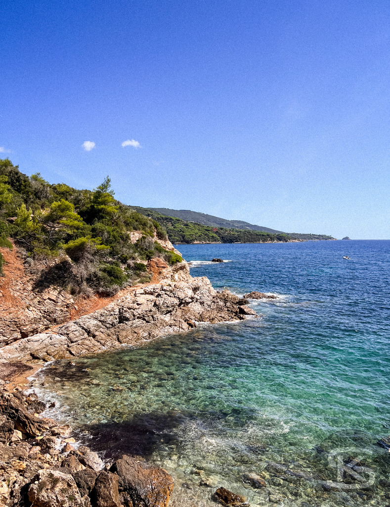 Kristallklare Buchten: Wilde Felsküste auf Elba – Wandbild | Blick auf eine zerklüftete Felsküste auf der Insel Elba; im Vordergrund kristallklares, türkisfarbenes Wasser, das in tiefes Blau übergeht, gesäumt von grüner mediterraner Macchia unter strahlend blauem Himmel. - Realisiert mit Pictrs.com