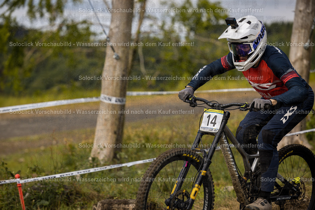 Action im Bikepark Winterberg | Ein Mountainbiker rast mit seinem Specialized Mountainbike durch den Bikepark Winterberg. Er trägt eine Nummer 14 und bereitet sich auf einen Sprung vor. Die Umgebung ist staubig und von Bäumen und Gras umgeben. Die Sonne scheint und sorgt für gute Sichtverhältnisse. - Realisiert mit Pictrs.com