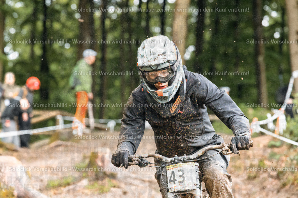 Mountainbiker im Wald | Ein Mountainbiker rast mit hoher Geschwindigkeit durch einen verschneiten Wald. Er trägt einen schwarzen Helm mit Visier und eine schwarze Jacke. Er trägt die Nummer 43 auf seinem Fahrradrahmen. Der Hintergrund zeigt verschneite Bäume und andere Mountainbiker. - Realisiert mit Pictrs.com