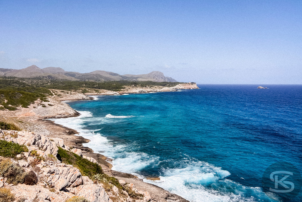Spektakuläre Steilküste und Panoramablick bei Cala Mesquida, Mallorca | Panorama-Aufnahme einer felsigen Steilküste auf Mallorca bei Cala Mesquida mit schäumender Brandung am Ufer, tiefblauem Meer und einer weitläufigen Hügellandschaft unter klarem Himmel. - Realisiert mit Pictrs.com