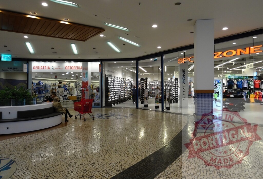 Shopping mall interior with retail stores and seating | Shopping mall interior featuring storefronts, patterned tiled floor, and a person sitting on a bench - Realized with Pictrs.com