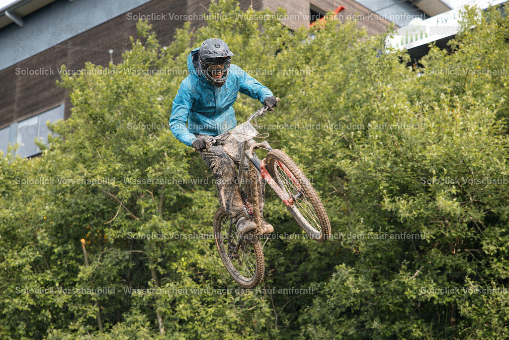 Action im Bikepark Winterberg | Ein Mountainbiker in blauer Regenkleidung springt in der Luft über eine dichte Hecke. Er trägt einen Helm und Handschuhe und sein Mountainbike ist stark verschmutzt. Im Hintergrund ist ein modernes Gebäude zu sehen. Die Szene spielt im Bikepark Winterberg, im Rahmen des iXS Downhill Cups 2025, speziell auf der Strecke 'The Mother'. - Realisiert mit Pictrs.com