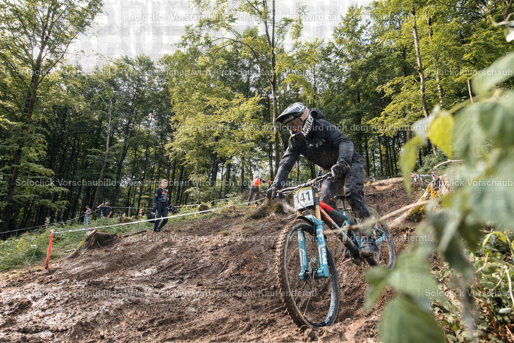 Mountainbike-Abenteuer im Wald | Ein Mountainbiker in voller Ausrüstung rast einen staubigen, kurvigen Pfad in einem dichten Wald hinunter. Er trägt einen Helm, eine Brille und eine Weste und fährt ein blaues Mountainbike mit roten Akzenten. Im Hintergrund sind weitere Mountainbiker und ein staubiger Weg zu sehen. - Realisiert mit Pictrs.com