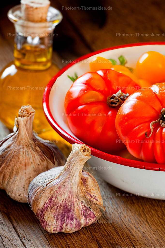 Mediterrane Küche: Tomaten, Knoblauch und goldenes Öl | Eine emaillierte Schüssel zeigt glänzende rote Tomaten neben frischen Knoblauchzehen auf rustikalem Holztisch. Eine kleine Glasflasche mit goldenem Olivenöl vervollständigt diese mediterrane Komposition. Das warme Licht betont die natürlichen Farben und Texturen der frischen Zutaten perfekt. - Realisiert mit Pictrs.com