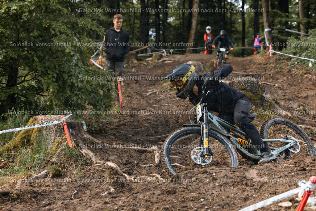 Mountainbike-Action im Schnee | Ein Mountainbiker in voller Ausrüstung rast einen steilen, verschneiten Hang hinunter. Er ist in einer dynamischen Position, die darauf hindeutet, dass er einen Sprung oder eine Abfahrt bewältigt. Der Hintergrund zeigt einen bewaldeten Hang mit weiteren Mountainbikern und Zuschauer. - Realisiert mit Pictrs.com