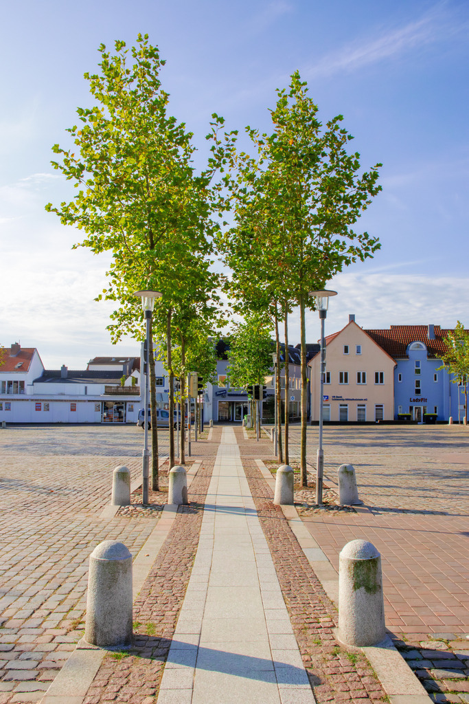 Wandbild: Kappeln erwacht – Ein stiller Moment am Morgen | Ein malerischer Frühlingsmorgen in Kappeln an der Schlei – dieses Wandbild zeigt einen ruhigen Weg, der in die Fußgängerzone führt. Die Bäume säumen den Weg mit frischem Grün, während die klare Morgenluft eine friedliche Atmosphäre schafft. Der fast leere Parkplatz verstärkt das Gefühl von Ruhe und Weite, bevor die Stadt zum Leben erwacht. Perfekt für eine stilvolle Raumgestaltung, die die Gelassenheit eines Morgenspaziergangs einfängt. - Realisiert mit Pictrs.com