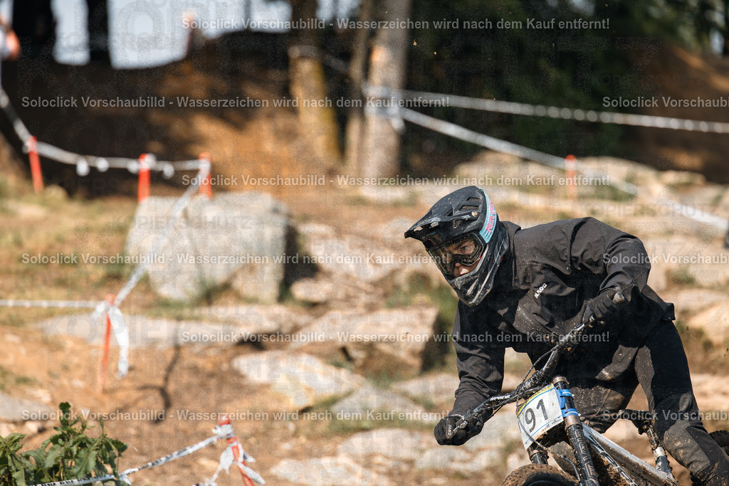 Mountainbiker im Full Speed | Das Bild zeigt einen Mountainbiker in voller Montur, der einen staubigen, felsigen Kurs durchquert. Er trägt einen schwarzen Helm mit Visier, eine schwarze Schutzbrille, eine schwarze Jacke und Handschuhe. Auf seinem Rücken befindet sich eine Nummer 91. Er fährt ein Mountainbike mit Federgabel und breiten Reifen. Der Kurs ist mit roten und weißen Markierungen versehen. Im Hintergrund sind Bäume und Felsen zu sehen. - Realisiert mit Pictrs.com