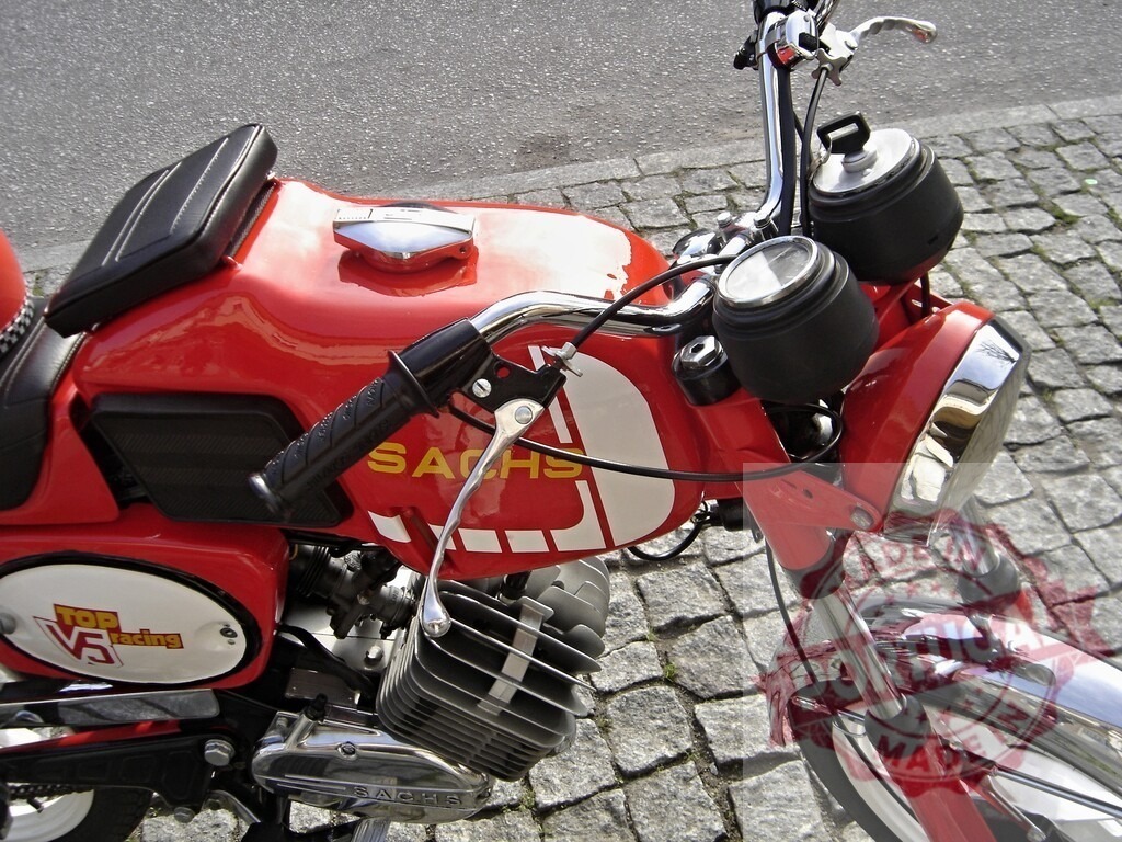 Red Sachs Motorcycle Parked on Cobblestone Street | Shiny red Sachs motorcycle with black seat and chrome details parked on a cobblestone street - Realized with Pictrs.com
